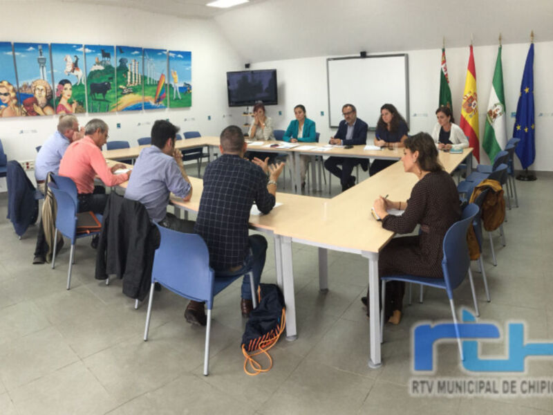 En una sala de reuniones, varias personas se sientan en mesas redondas. En la pared hay carteles con imágenes de playas y actividades, junto a banderas españolas y europeas. Una persona toma notas en un bloc de notas.