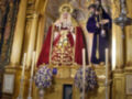 Altar de una iglesia con una imagen de la Virgen María vestida en rojo y dorado, rodeada por velas y flores. Arriba hay un arco dorado con detalles geométricos. A la derecha, una figura vestida en azul con un sombrero y un bastón. El entorno es dorado y ornamental, con detalles de madera y cristal.