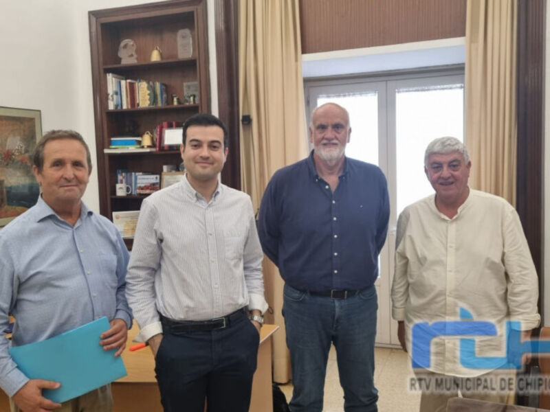 Un grupo de cuatro personas posa frente a una estantería llena de libros y objetos decorativos. La imagen muestra un ambiente formal, posiblemente en una oficina o sala de reuniones. La luz natural entra a través de las ventanas, iluminando el espacio y creando un ambiente acogedor. La presencia de una carpeta azul en la mano del individuo a la izquierda añade un toque de profesionalismo. En el fondo, se observa una pintura colgada en la pared y un cuadro sobre la mesa. La imagen está fir...