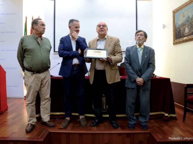 Un grupo de hombres posa en un escenario formal, uno sostiene un premio mientras los otros observan. El ambiente es elegante con una pared de fondo que incluye un lienzo y una bandera. La iluminación es adecuada para la celebración.