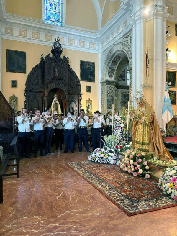 Imagen de una iglesia con un coro de músicos tocando instrumentos tradicionales. La arquitectura es elegante con detalles dorados y una estatua de la Virgen en el centro. El suelo es de mármol y hay flores blancas y rosadas alrededor.