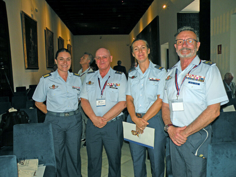 Una imagen de cuatro personas en uniforme militar, posando con sonrisas. La escena parece ser en un lugar formal o ceremonial, con sillas y carteles de fondo.