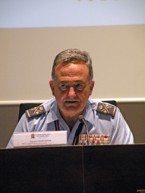Un hombre en uniforme militar se encuentra frente a un micrófono, probablemente dando una conferencia o discurso. La imagen muestra detalles de su uniforme y un nombre plateado en el fondo, pero no se puede leer claramente.