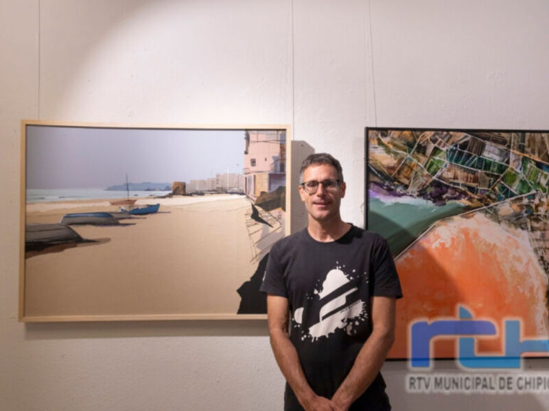 Un hombre con una camiseta negra posa frente a dos cuadros de playa. Uno muestra una imagen de la playa con casas y el mar, mientras que el otro presenta un mapa de Chipiona. La imagen se encuentra en una pared blanca con la marca "RTV MUNICIPAL DE CHIPIONA" en la esquina inferior derecha.