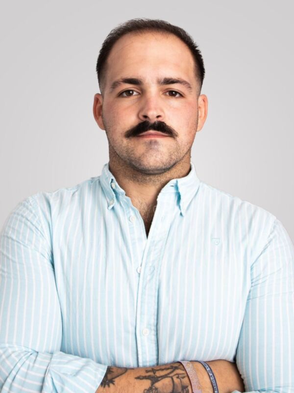 Un hombre con una barba y bigote, vistiendo una camisa de manga larga a rayas azules. Sus brazos están cruzados en el pecho, y se puede ver un tatuaje en su muñeca izquierda. El fondo es de color claro, probablemente gris o blanco.