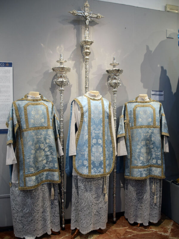 Imagen de tres vestiduras religiosas, posiblemente para un servicio eclesiástico. Las vestiduras son de color azul claro con detalles dorados y bordados, y están acompañadas por dos crucifijos de metal. La escena se encuentra en un espacio interior, posiblemente una sala o museo, con un fondo de pared gris y suelo de baldosas marrones.