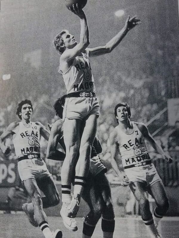 Un jugador de baloncesto en acción, levantado sobre sus compañeros durante un partido. El jugador lleva una camiseta con el nombre "REALE" y la marca "MARLON", mientras que sus compañeros también llevan camisetas con el nombre "REALE" y la marca "MARLON". La escena se desarrolla en un estadio lleno de espectadores.