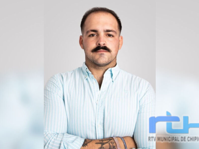 Un hombre con barba y bigote, cabello corto y rasgado, mirando hacia la cámara. Está vestido con una camisa de manga corta a rayas azules y blancas, con los brazos cruzados en frente. El fondo es gris claro, destacando su rostro y la camisa. En la parte inferior derecha de la imagen hay un logotipo azul y blanco con el texto "RTV MUNICIPAL DE CHIPIONA".