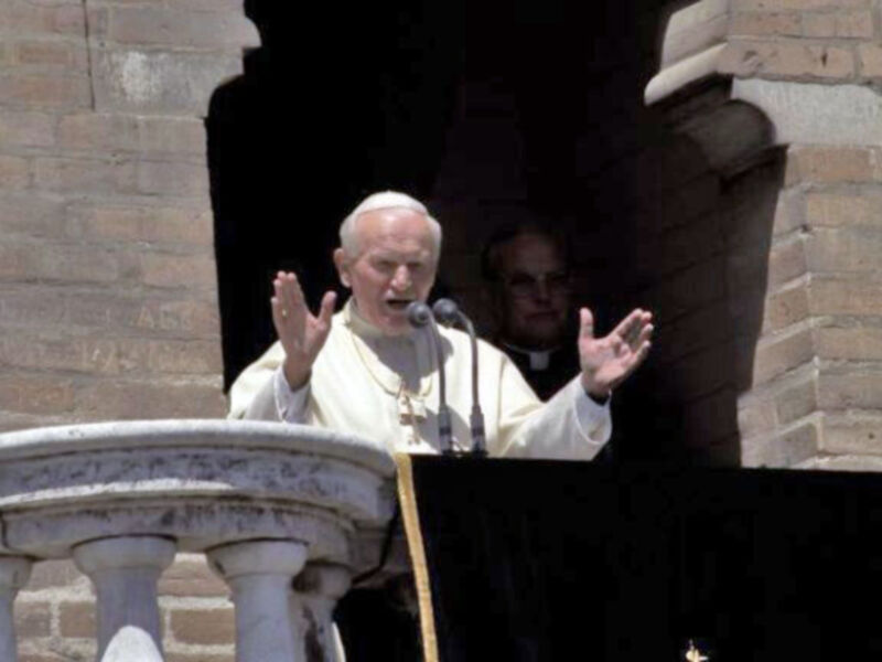 El Papa Benedicto XVI pronuncia una oración desde un balcón, con manos extendidas y gestos expresivos. La imagen muestra una arquitectura de estilo barroco, probablemente en el Vaticano.