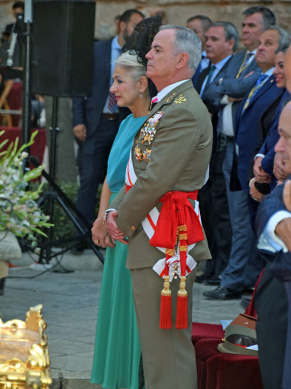 Imagen de una ceremonia militar donde un oficial en uniforme de gala y una mujer vestida formalmente están presentes. El fondo muestra un edificio histórico con columnas y una granja de flores. La escena sugiere una celebración o evento oficial, posiblemente militar.