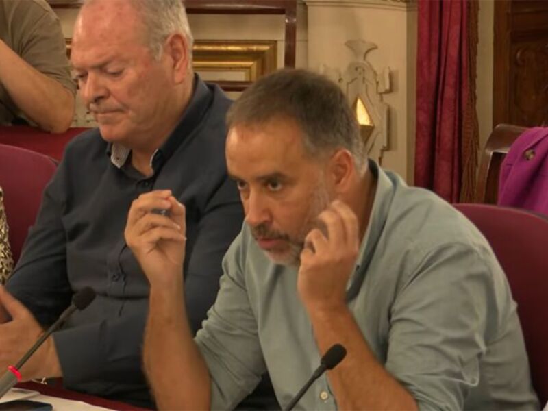 Un hombre en un traje oscuro habla mientras otros asistentes escuchan atentamente. La imagen muestra una reunión formal con micrófonos en mesa y personas sentadas en sillas rojas.