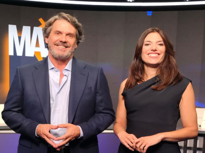 En la imagen, dos presentadores se encuentran en un estudio de televisión. El hombre está vestido con una camisa blanca y corbata, mientras que la mujer lleva un vestido negro. Ambos sonríen y tienen una expresión amistosa, lo que sugiere un ambiente relajado y agradable. La escena está ambientada en un estudio con una pantalla de fondo que muestra la palabra "MA".
