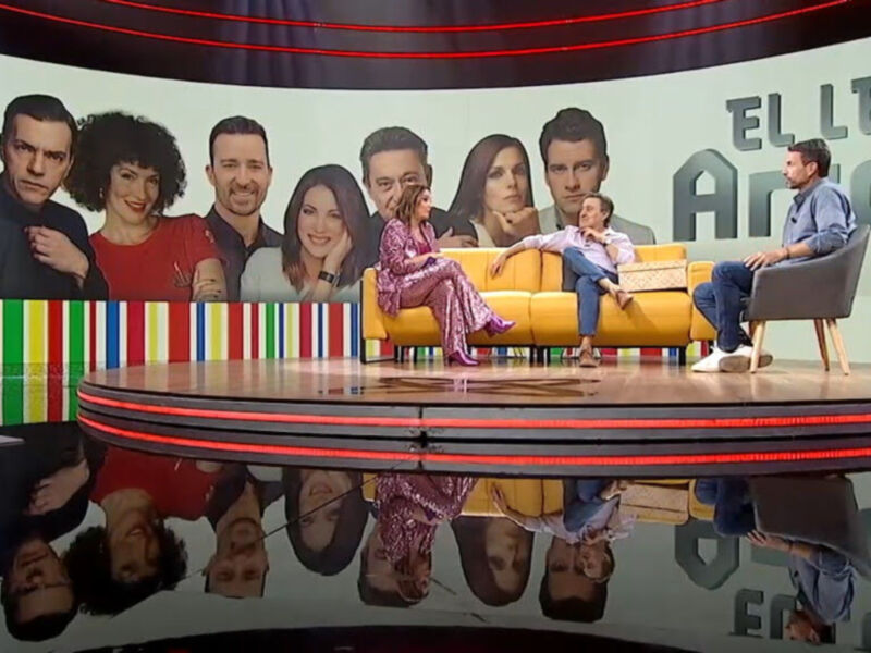 Una escena de un programa de televisión con una mesa redonda en el centro, donde varios presentadores y invitados se sientan. La pared de fondo tiene un diseño gráfico con el texto "EL LEGADO". El programa parece ser una conversación o entrevista.