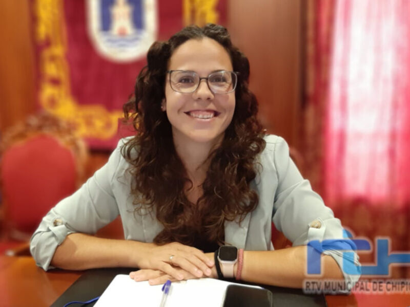 A una mujer sentada en un escritorio, sonriendo y mirando hacia la cámara. El fondo incluye una bandera roja con un escudo dorado y letras blancas, junto a cortinas rosadas. La imagen es parte de una publicación oficial con el logotipo y nombre del municipio en la esquina inferior derecha.