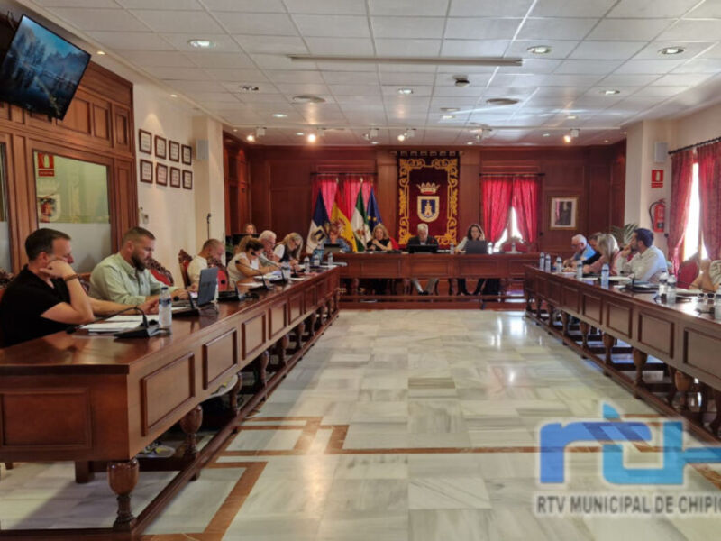 Sala de reuniones con mesa central, sillas para los asistentes y un televisor en la pared. La sala está decorada con banderas y un escudo heráldico.