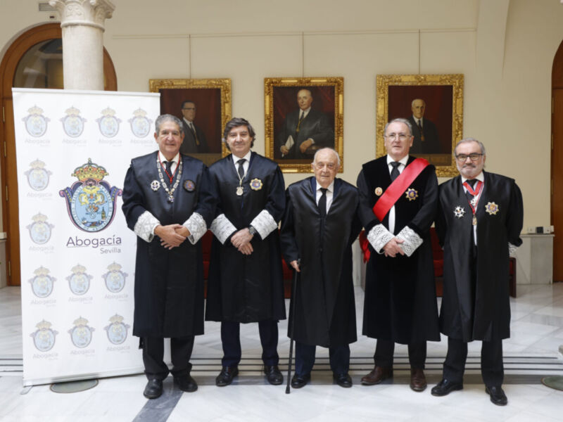 Fotografía de un grupo de hombres vestidos formalmente, posando frente a una gran bandera con insignias y el texto "Abogacía". El fondo muestra cuadros de retratos históricos.
