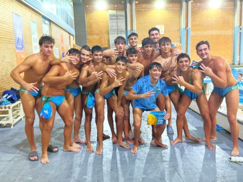 Un grupo de jóvenes nadadores posa en la orilla de una piscina, mostrando sus cuerpos musculosos y sonrientes. Algunos llevan trajes de baño azules, mientras que otros usan camisetas deportivas. La escena se desarrolla en un ambiente acuático, con el agua visible al fondo y una pared de azulejos amarillos.