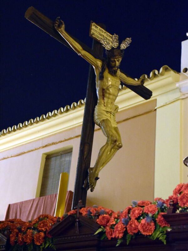 Cristo crucificado en una cruz dorada, con flores de color naranja y rosa al pie. Fondo nocturno con edificio amarillo en la perspectiva.