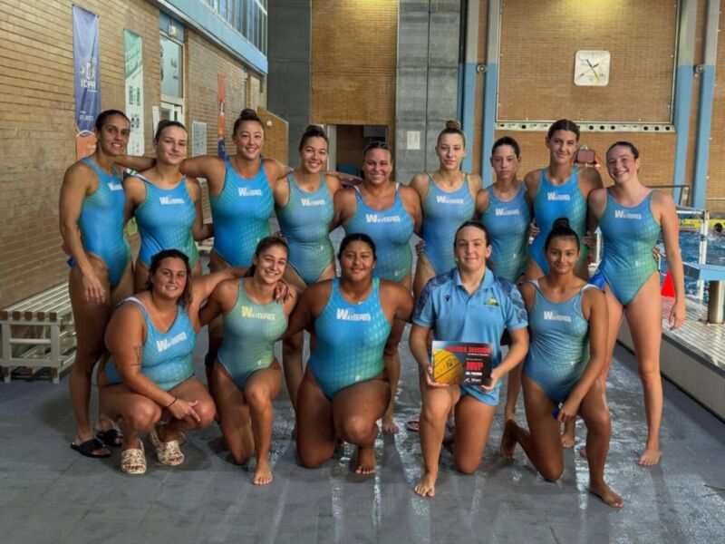 Equipo de natación femenina posando en la piscina. Todos están vestidos con trajes de baño azules y blancos, y una mujer del centro sostiene un trofeo. La imagen se toma en un gimnasio con bancales y una pared de azulejos.