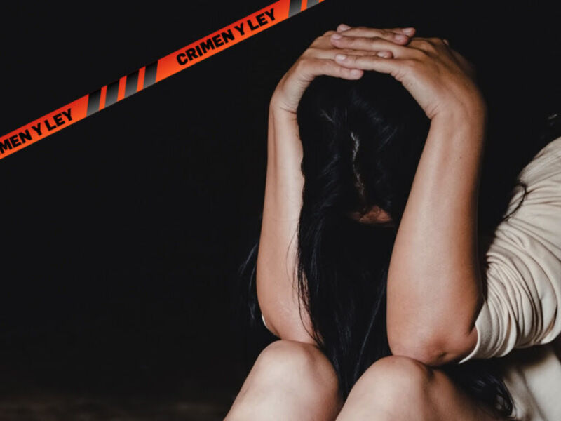 Una mujer con cabello oscuro se sienta con las piernas cruzadas, cubriendo su rostro con sus manos. En el fondo, hay un cinta de seguridad con la palabra "CRIMEN Y LEY" en rojo. La escena parece ser nocturna o en un lugar oscuro.