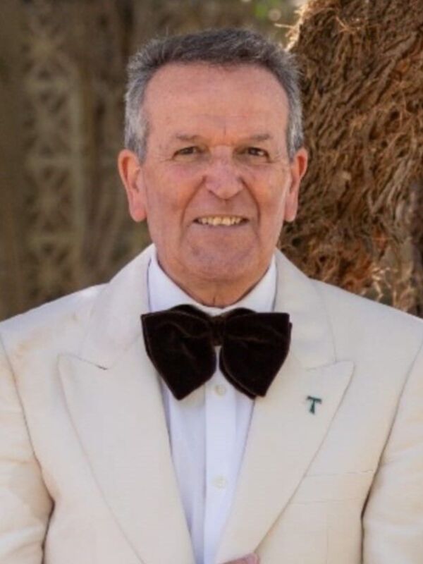 Un hombre con una camisa blanca, corbata de terciopelo negro y un chaleco beige. Detalle: una insignia con la letra "T" en el pecho. Fondo: un árbol y una estructura de madera.