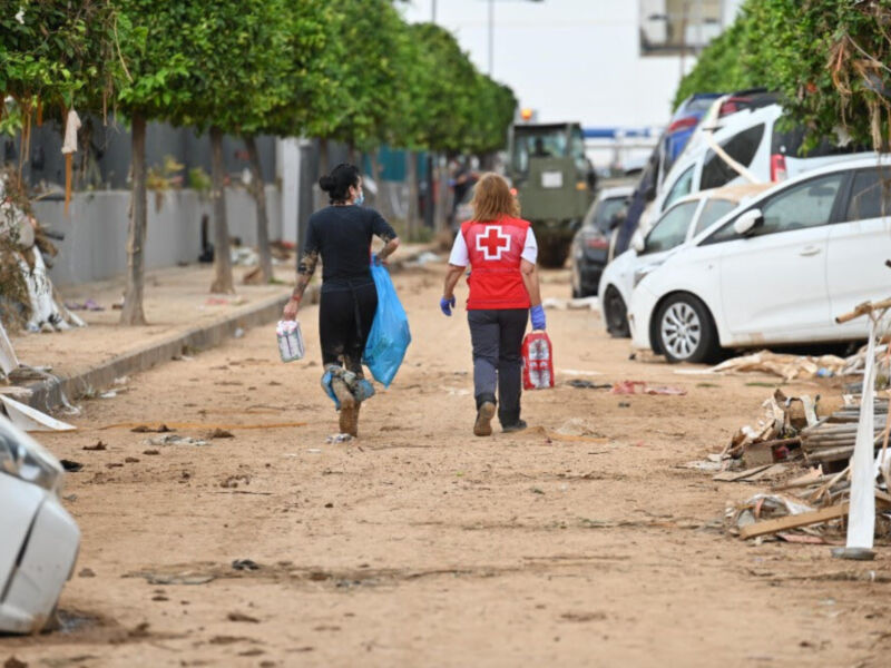 Dos personas caminan por un camino desolado, uno con una mochila y otro con un saco de plástico. La escena muestra evidencias de desastre, con escombros y basura alrededor.