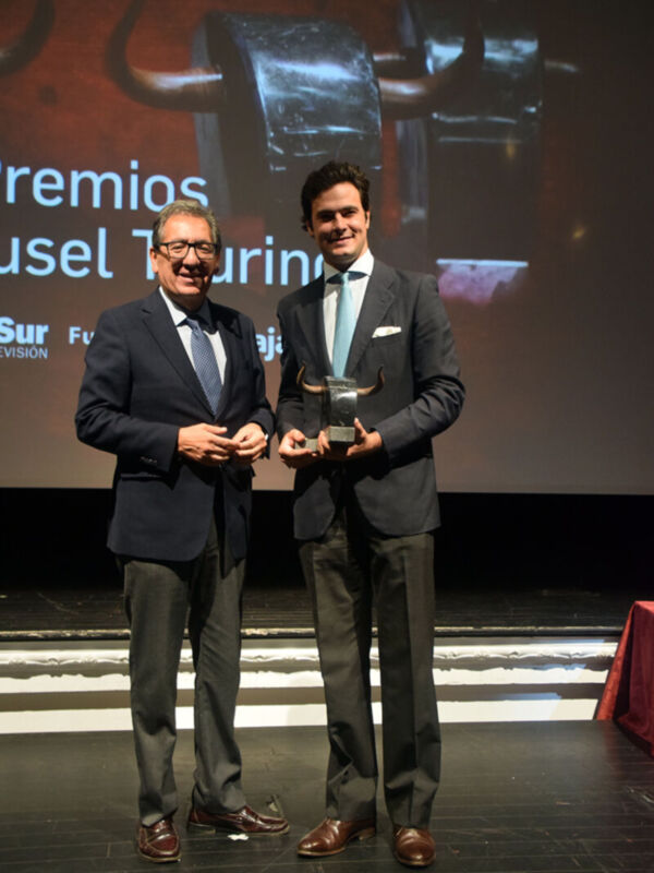 Dos hombres en trajes posan con un premio, frente a una pantalla que muestra imágenes de vehículos.