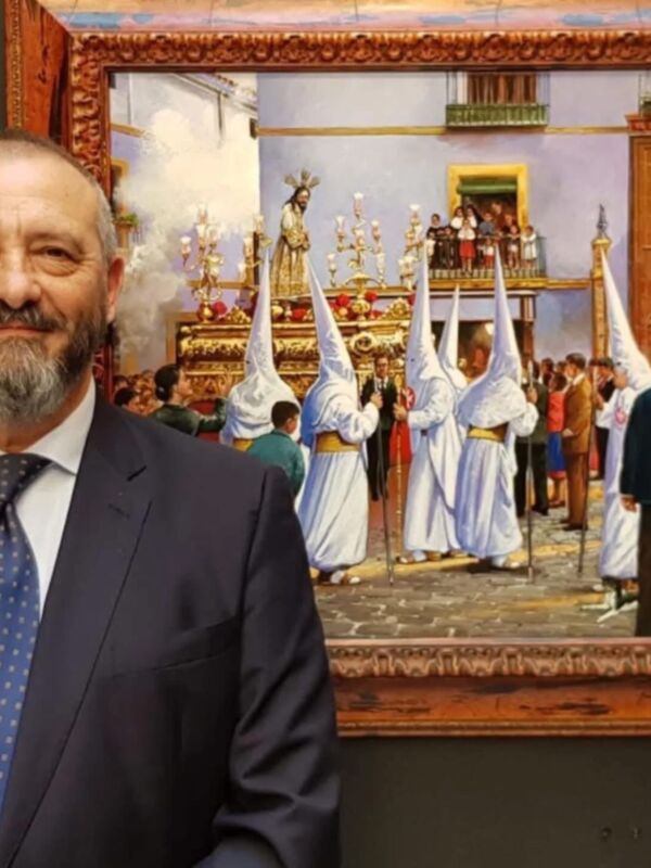 Un hombre con barba y traje azul posa frente a una pintura que muestra una procesión religiosa con personajes vestidos de blanco y azul. La escena se desarrolla en un entorno histórico con detalles arquitectónicos y decorativos.