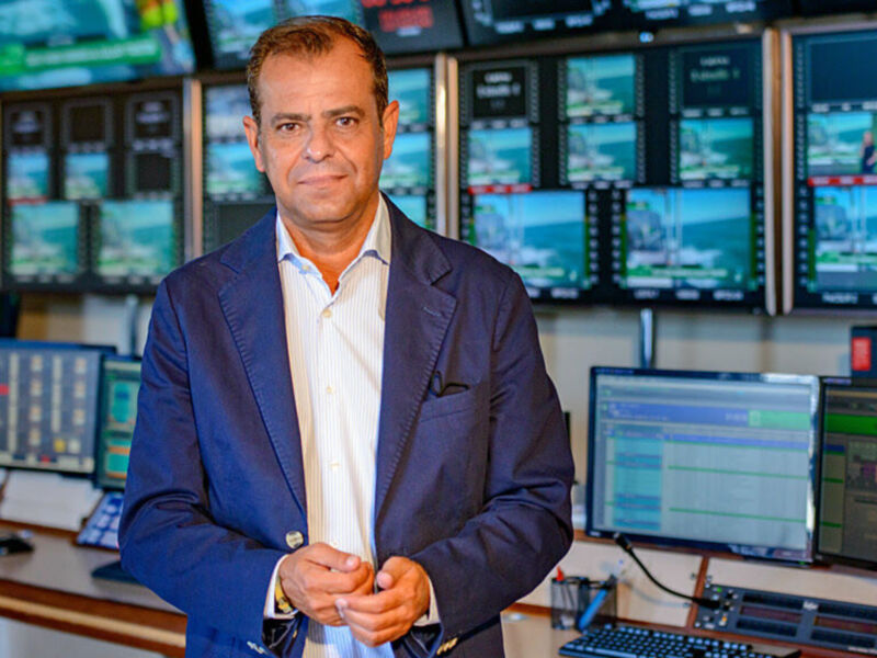 Un hombre en una oficina de control, con varios monitores en el fondo. El ambiente parece ser un lugar de trabajo tecnológico o de control.