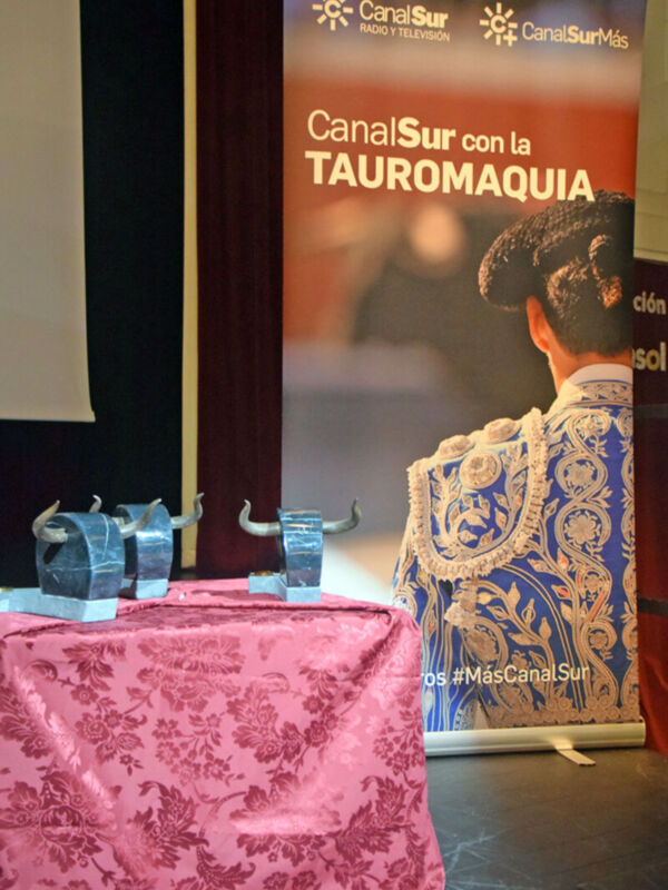 Se muestra una mesa cubierta con un mantel rosa y dos trofeos de toro en el centro. En el fondo, hay un panel con la publicidad de Canal Sur y la frase "CanalSur con la TAUROMAQUÍA".