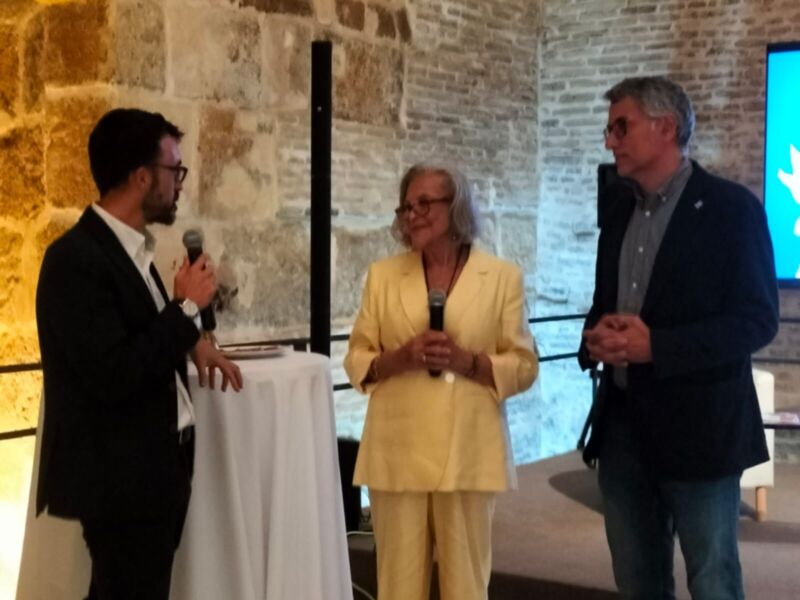 Tres personas en un evento formal, dos hombres y una mujer, con micros en mano. La imagen muestra una pared de ladrillo y un fondo con pantallas.
