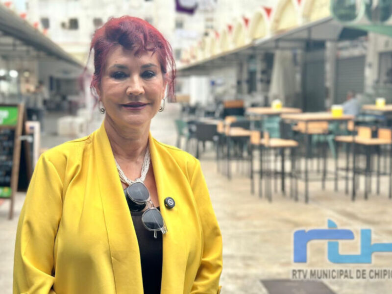 A una mujer con cabello rojo y gafas de sol en la oreja, vestida con un abrigo amarillo. Se encuentra en una zona con mesas y sillas, posiblemente un espacio público o comunitario. En la parte inferior derecha de la imagen, hay una marca con el texto "RTV MUNICIPAL DE CHIPIONA".