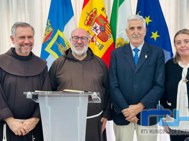 En la imagen, se observan cuatro personas frente a una bandera española y otras de diferentes países. Dos hombres vestidos con hábitos religiosos se encuentran a la izquierda, mientras que dos personas más, uno de ellos con una corbata azul y la otra con un uniforme formal, se encuentran a la derecha. La escena parece ser una reunión oficial o un evento institucional, posiblemente relacionado con la administración local de Utrera.