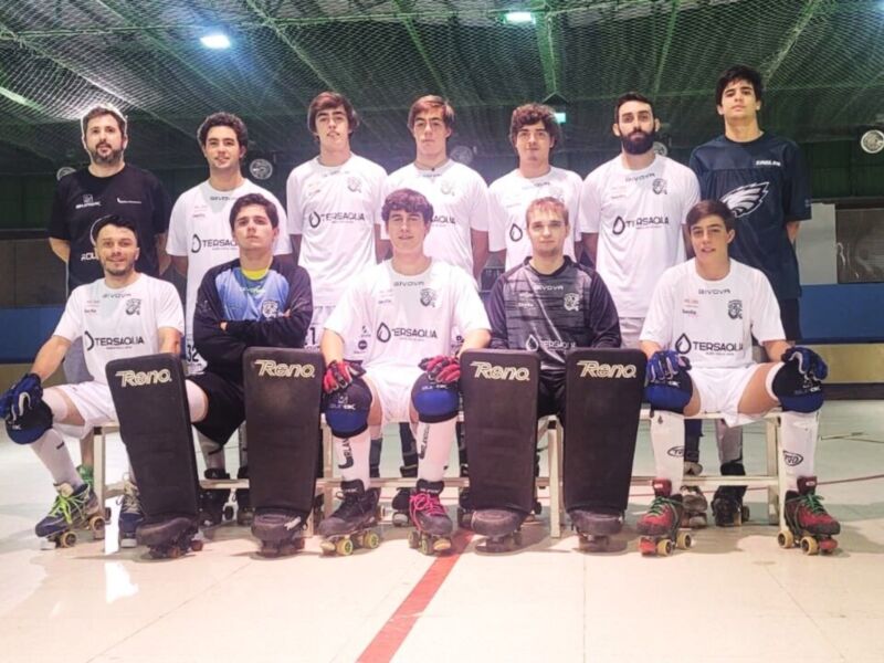 Equipo de hockey sobre patines en un gimnasio. Los jugadores están sentados con sus patines, listos para el juego. El equipo lleva camisetas blancas y azules con logos, y patines de color rojo. La iluminación es brillante y la superficie del gimnasio es de color claro, probablemente azul.