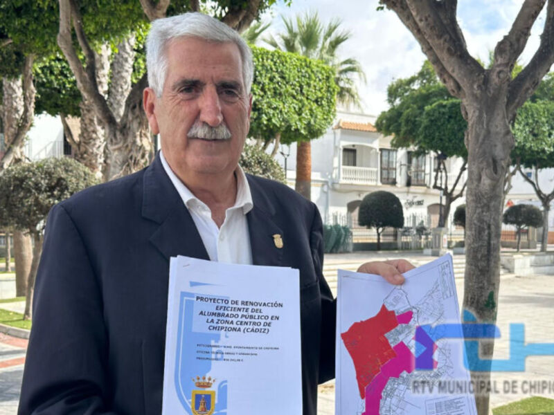 Un hombre con barba blanca sostiene dos folletos. El fondo muestra un parque con árboles y una estructura urbana en el horizonte. La imagen es de Chipiona, Andalucía, España.
