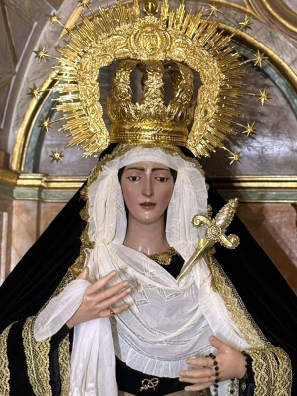 Una estatua religiosa de una Virgen María vestida con un manto negro y una saya blanca, adornada con detalles dorados. La estatua está coronada con un halo de estrellas y una corona dorada, lo que la hace parecer majestuosa y sagrada. La escena se desarrolla en un entorno arquitectónico con columnas doradas y una bóveda, lo que sugiere un ambiente de iglesia o templo. La estatua parece estar en una posición de oración, con las manos juntas y el rostro sereno.