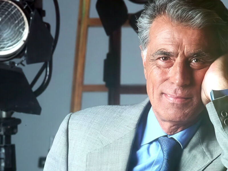 Un hombre con una camisa azul y corbata posa frente a un estudio de cine, con una cámara en el fondo.