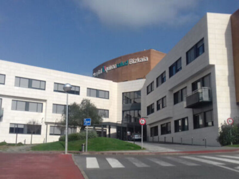 Edificio moderno con fachada blanca y roja, detalles en negro. Sobre él, se lee "Quintal Tech Bizkaia". A la izquierda, árboles y vegetación. En el frente, se ven señales de tráfico y un poste con luz. Sobre la imagen, cielo azul con algunas nubes.