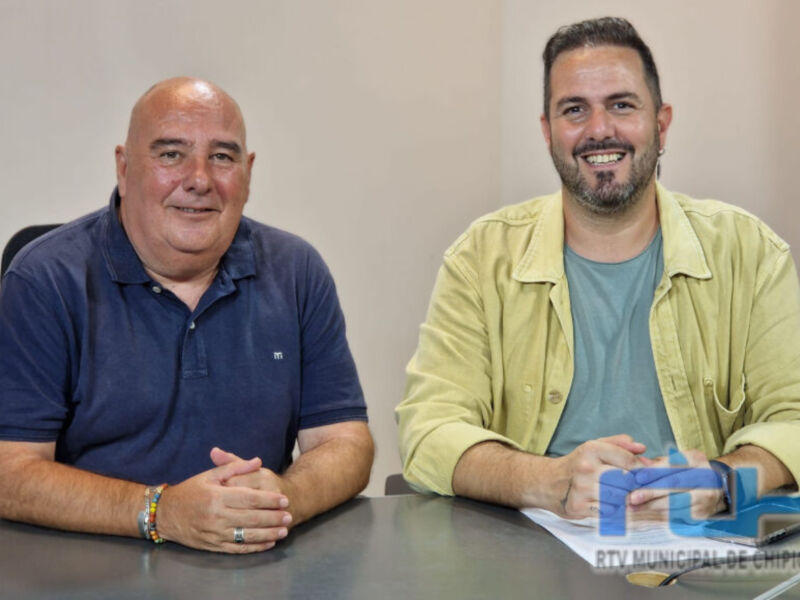 Dos hombres sentados en una mesa, uno con camisa azul y el otro con camisa amarilla. Ambos sonríen ampliamente. En la mesa hay documentos y una computadora portátil. La imagen parece ser de una reunión o entrevista formal.