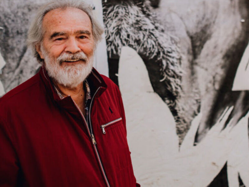 Un hombre con barba blanca y cabello gris, vestido con una chaqueta roja, posa frente a un fondo blanco con imágenes de elefantes.