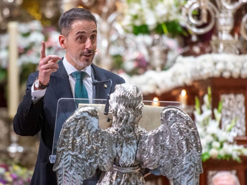 Un hombre en traje negro sostiene un libro abierto, frente a una estatua de ángel con alas doradas y flores blancas en el fondo.