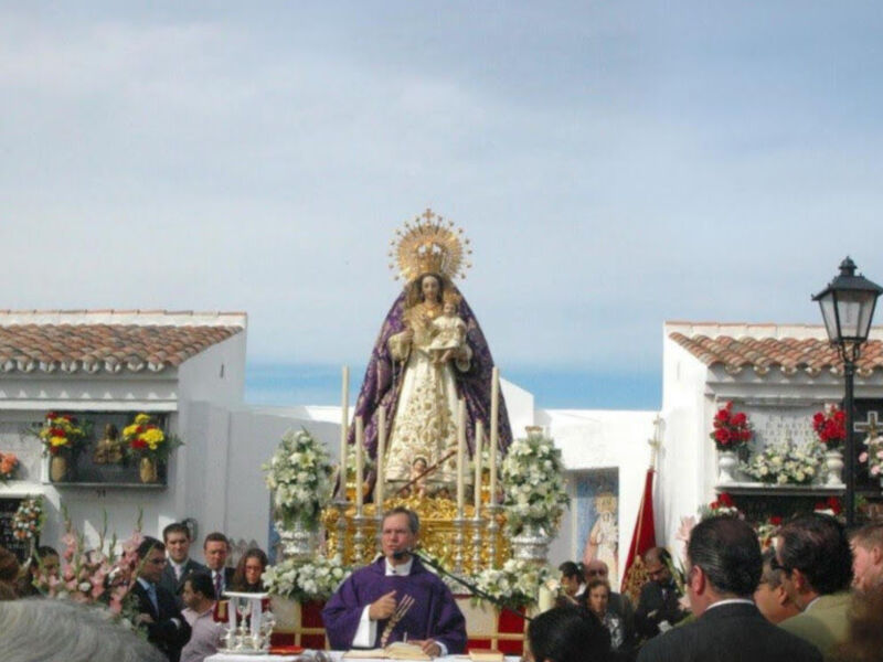Una procesión religiosa con una estatua de la Virgen María en el centro, rodeada por flores y decoraciones. El cielo es azul con algunas nubes. La imagen muestra a personas vestidas formalmente, incluyendo un sacerdote en el centro. La escena se desarrolla al aire libre, con edificios y una farola visible en el fondo.
