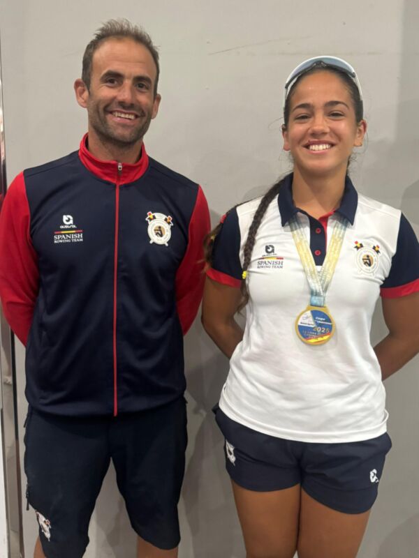 Dos atletas posan juntos, uno con una medalla dorada en la cintura. Ambos lucen uniformes deportivos de color azul y blanco, con detalles en rojo. La imagen sugiere un logro deportivo importante.