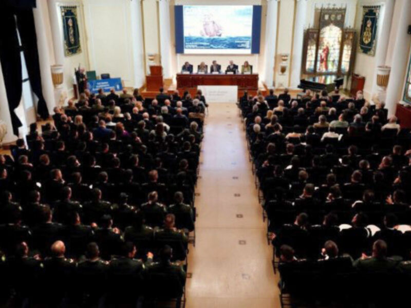 Una sala de conferencias llena de personas sentadas en filas, con una pantalla grande en el fondo mostrando un dibujo. La sala está decorada con carteles y una gran ventana en el lado derecho.
