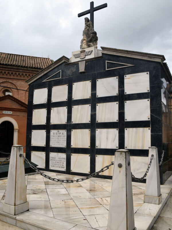 Una estatua de un hombre con una cruz en la cabeza se encuentra sobre un monumento negro y blanco, rodeado de una cadena. La estatua está en la parte superior del monumento, que tiene placas de mármol blancas. El monumento está en una plaza con un edificio rojo y otras estructuras alrededor. El cielo es nublado.