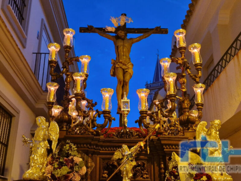 Cristo crucificado en una procesión religiosa, iluminado por faroles y ángeles dorados. En el fondo, una iglesia con detalles arquitectónicos.