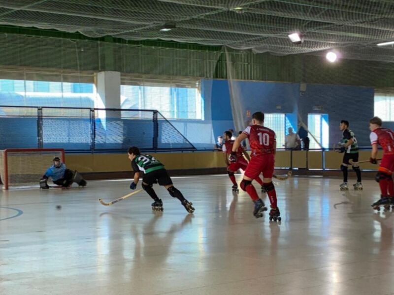 Un grupo de niños juega hockey en una cancha interior. El suelo es de baldosas y el techo tiene luces. Algunos niños llevan cascos, mientras otros no. La portería está en el extremo izquierdo de la imagen.