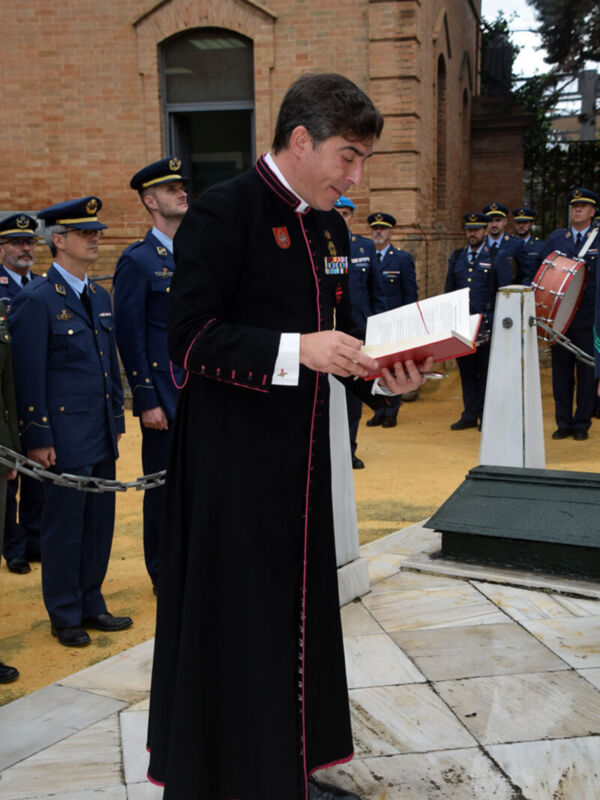 Un hombre en uniforme de gala negro con detalles rojos sostiene un sobre abierto. Está en una zona ceremonial, rodeado de otros oficiales uniformados y un edificio de ladrillo en el fondo.