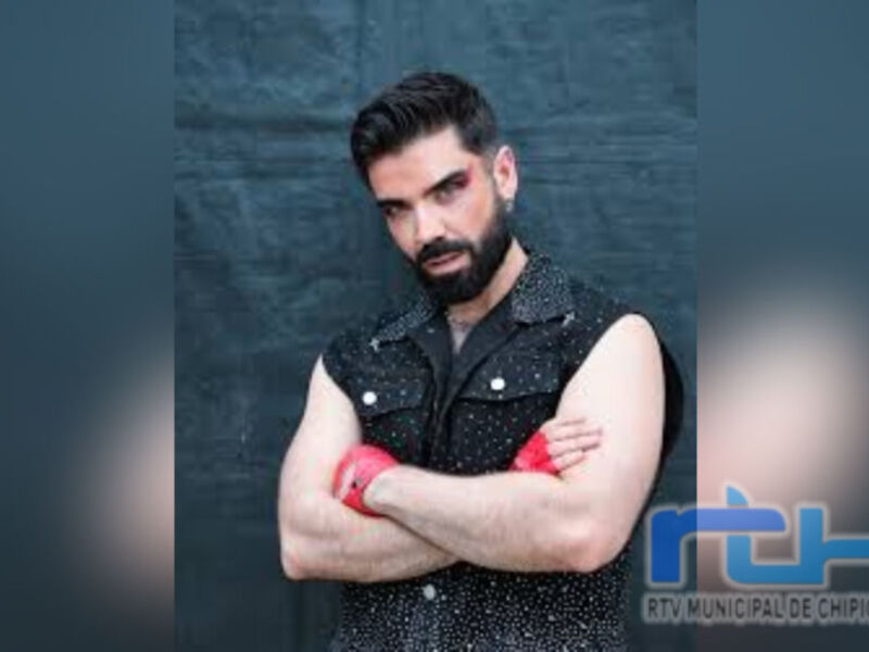El hombre con una barba y un vestido negro con detalles rojos, posando con los brazos cruzados frente a una pared gris.