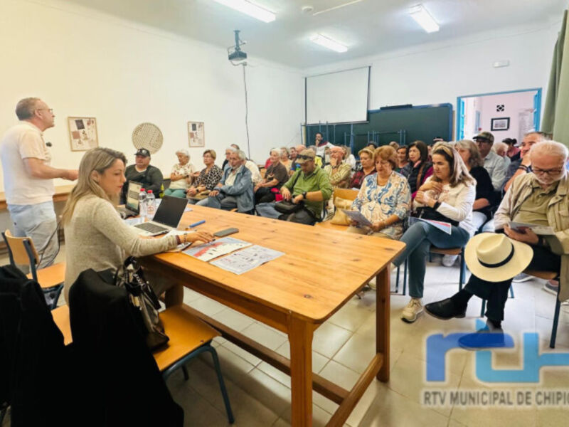 En una sala comunitaria, un grupo de personas asisten a una reunión informativa. La mesa central tiene un portátil y folletos, mientras que los asistentes observan atentamente. La imagen se sitúa en Chipiona, como lo indica el logotipo de la RTV Municipal.